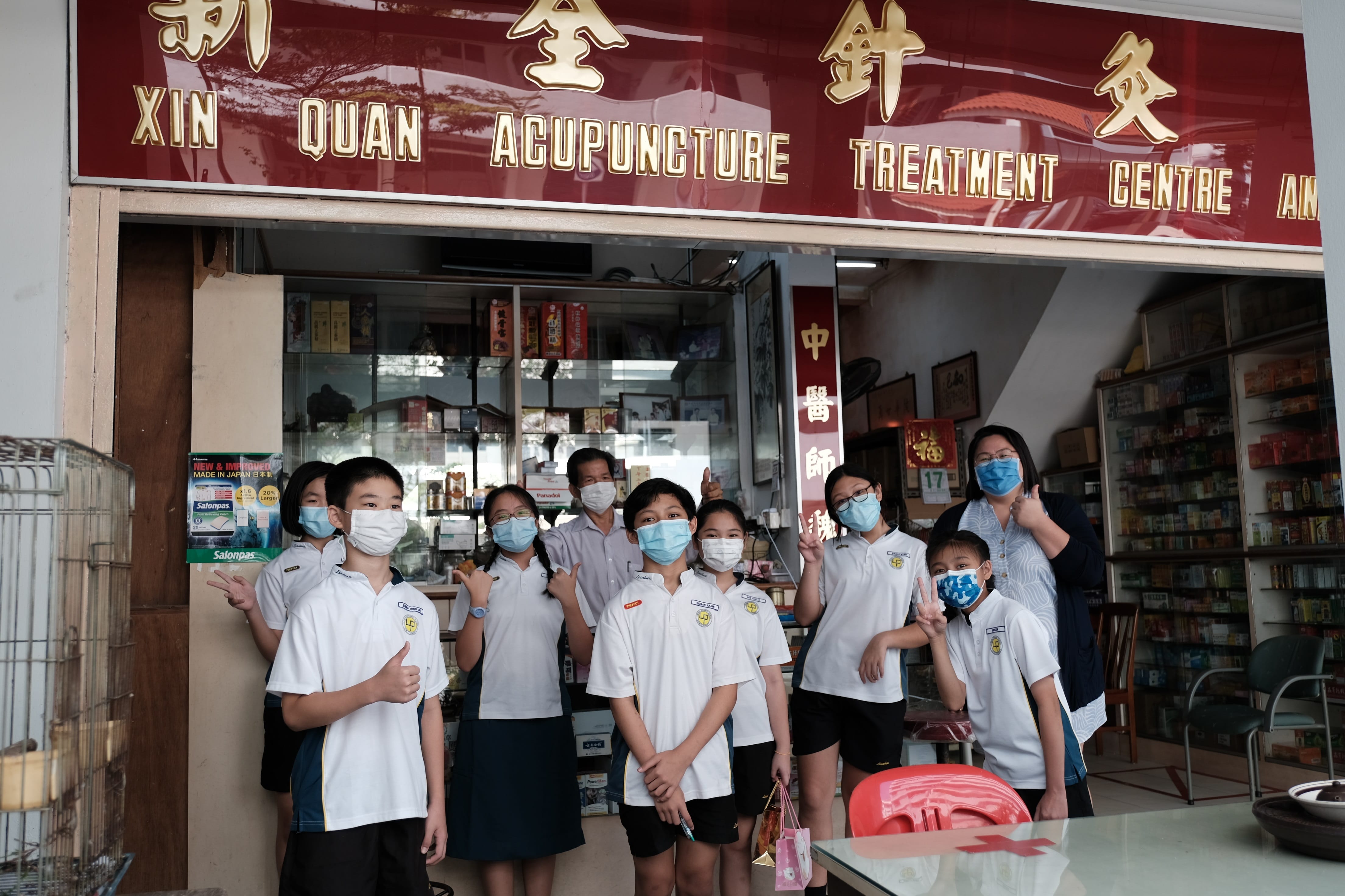 A group of masked children in school uniforms pose outside Xin Quan Acupuncture Treatment Centre with two adults.